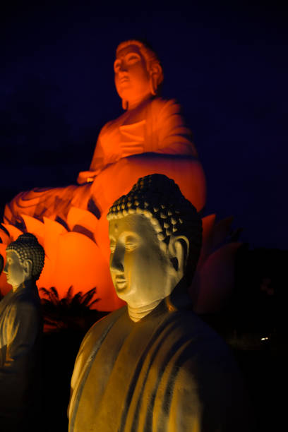 This captivating image depicts a large Buddha statue seated on a lotus flower, beautifully illuminated by orange and yellow lights against a dark background. The Buddha faces right, eyes closed, radiating peace and serenity in his traditional robe. In front, two smaller illuminated Buddha statues mirror this tranquil pose. The soft glow created by the lights enhances the spiritual atmosphere, making it a stunning night scene.