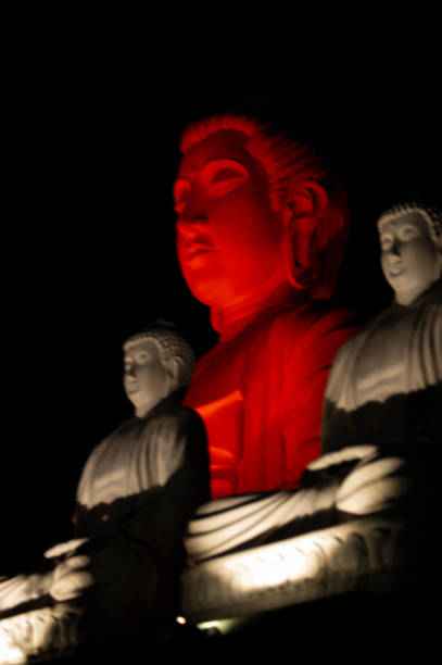 This captivating close-up image features a large Buddha statue illuminated with vibrant red and white lights, creating a stunning contrast against a dark background. With his head turned to the side and eyes closed, the Buddha wears a flowing red robe and displays a peaceful expression. Flanking him are two smaller Buddha statues, also aglow in red, all seated in tranquil meditative poses. Captured from a low angle, the composition emphasizes the statues' serene beauty.