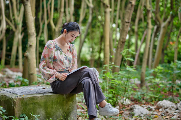 Side view of one asia young female reading book sitting on the stone bench at backyard's forest.