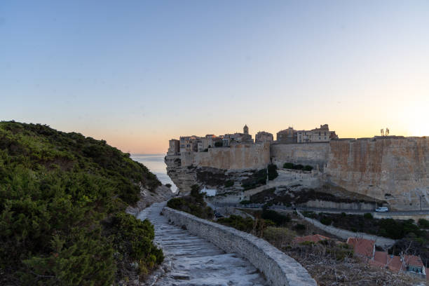 A cliffside village with ancient stone buildings at sunset, featuring a stone pathway leading to the town, set against a peaceful coastal and mountainous backdrop.