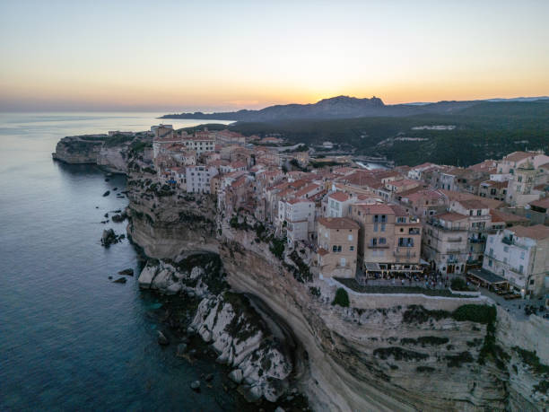 Aerial view of a historic cliffside town perched above the sea at sunset, with vibrant buildings cascading down the cliffs and soft evening light illuminating the horizon.