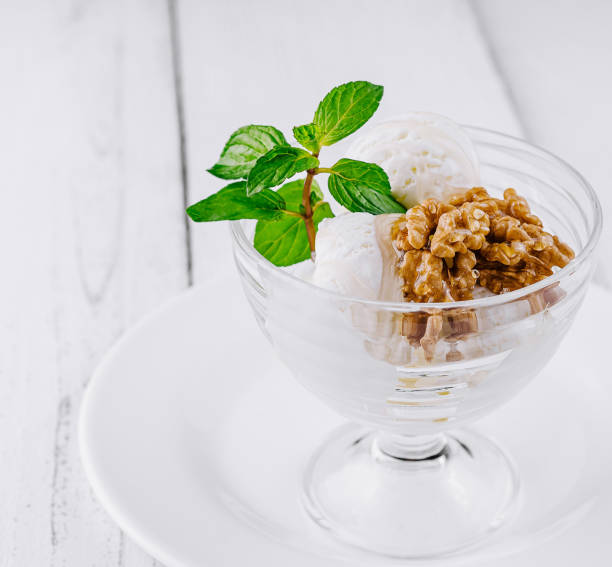 Glass cup presenting delicious vanilla ice cream with walnuts and mint leaves on white rustic background