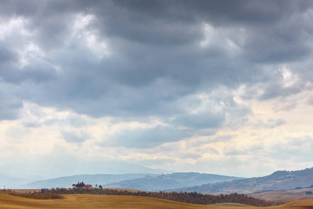 A moody landscape of Tuscany with dark, dramatic clouds looming over rolling hills. A lone farmhouse with cypress trees is visible in the distance, nestled among golden fields, creating a contrast between the earthy tones and the brooding sky.