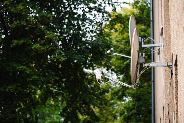 A satellite dish mounted on the side of a building, with a backdrop of lush green trees. The modern technology contrasts with the natural setting, highlighting the blend of urban infrastructure and nature.