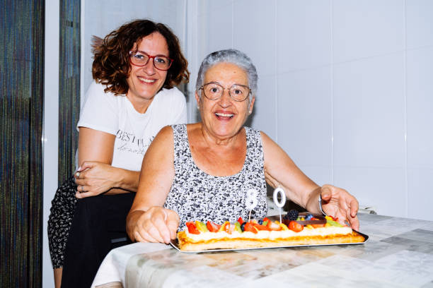 Portrait of Smiling Granny Celebrating Her 80th Birthday With Her Daughter and a Colorful Cake at Home A grandmother and her daughter pose together looking at camera, highlighting an 80th birthday cake adorned with candles and fruit. Their smiles capture the warmth of family celebration joyful moment. food recall stock pictures, royalty-free photos & images