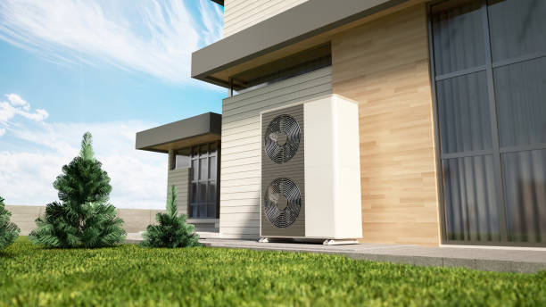 A heat pump is situated outside a modern home with wood siding. The unit is white with two fans and is positioned next to a window. Lush green grass covers the ground in front of the home.