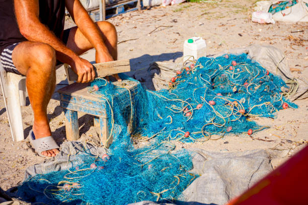 A fisherman sits on a chair at the beach and cleans fishing net from shells, smashing them with a wooden board.