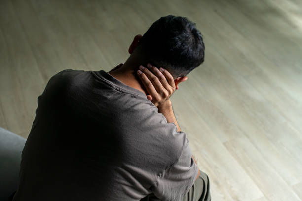 Medium close up image of a mid adult man in his 30s sitting on his bedroom floor at home. The man has a negative expression on his face, and appears depressed or as if he has received bad news.