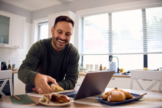 istock Happy man eating sandwich while working on laptop in the kitchen. 2176665908