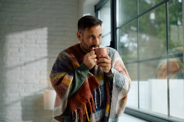 istock Man wrapped in a blanket drinking tea by the window. 2176666198