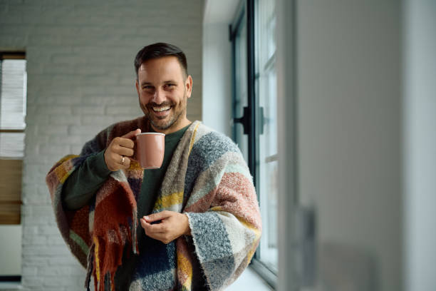 istock Happy man wrapped in blanket drinking tea and looking at camera. 2176666215