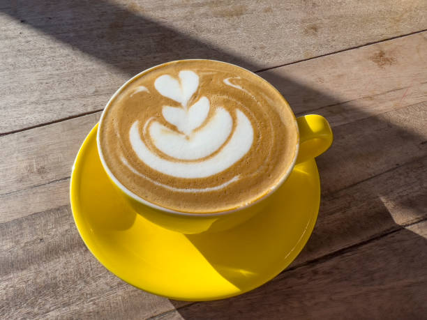 Close-up shot of a yellow coffee cup and saucer on a wooden table. The coffee has a pattern in the foam, and there is sunlight streaming across the frame.