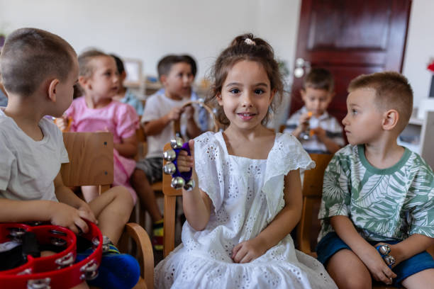A happy girl holds musical instruments in a classroom, enjoying a moment of learning and fun while surrounded by other children engaged in creative activities.