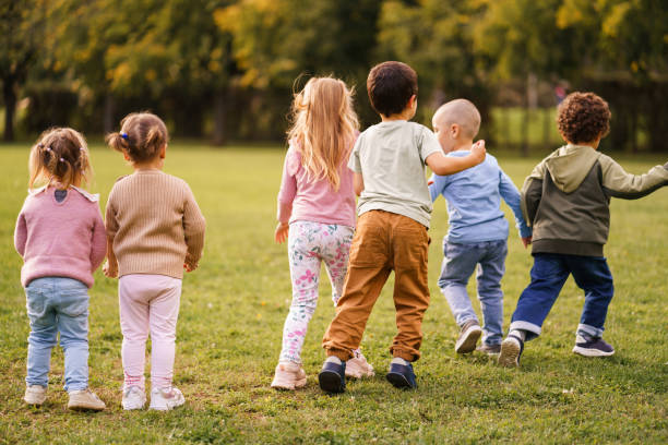 Group of multiethnic children and young Caucasian woman teacher walking together in green park, teacher leading children through grassy area surrounded by tall trees during outdoor activity