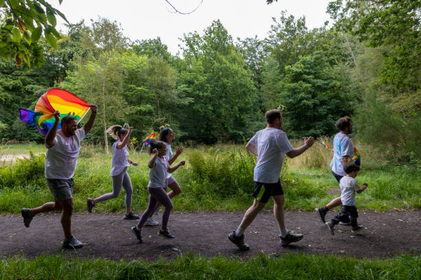Medium shot of two families running in an LGBTQ+ colour run in a woodland area in Gateshead. They are wearing white T-shirts and are covered in paint. The father of one of the families is running while carrying a rainbow flag.Videos are available similar to this scenario.