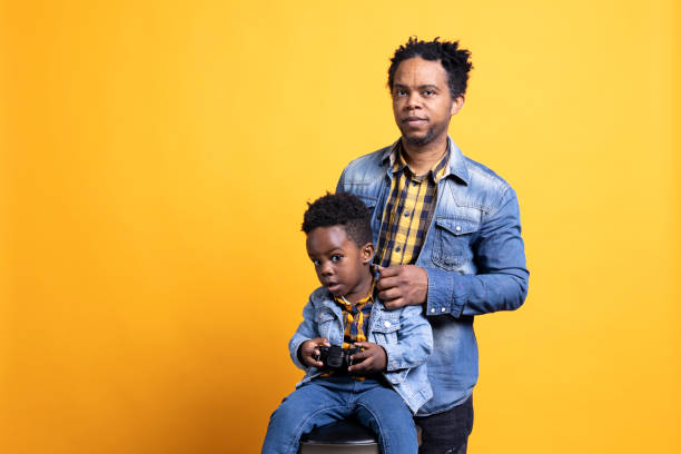 African american small boy playing video games on controller next to his father, posing against yellow background. Cheerful little child having fun with gaming online, sitting next to his parent.