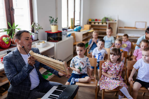 A teacher in a checked blazer is demonstrating how to use a percussion instrument while surrounded by young students sitting and observing in a classroom setting.