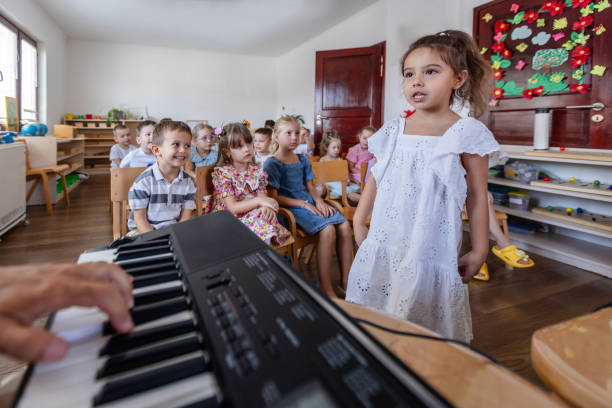 A young girl stands confidently at the front of a classroom, singing and performing while her peers sit in the background, creating a cheerful atmosphere.