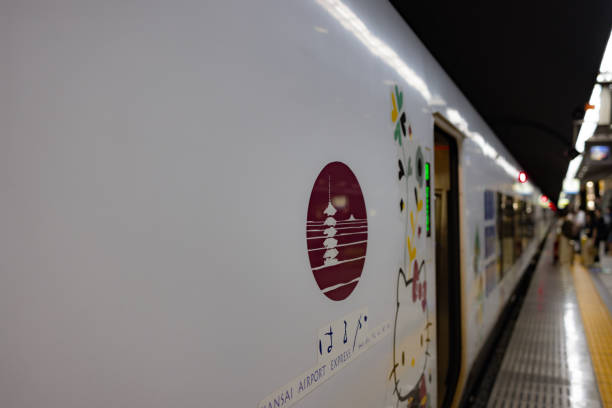 A sleek and modern Japanese train at a station platform, featuring a minimalist design with a maroon emblem and clean white exterior. The emblem displays a traditional Japanese motif, reflecting a blend of modern transportation and cultural aesthetics. A glimpse of commuters and platform details adds to the lively atmosphere.