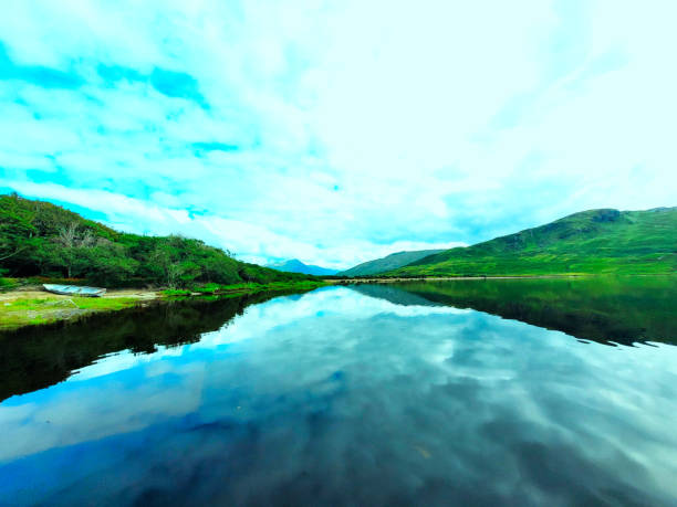 Kylemore Lough in Connemara, Ireland. The still waters perfectly reflect the passing clouds above, creating a mirror-like effect. A charming boat rests quietly on the shore, adding a touch of rustic charm to the picturesque landscape.