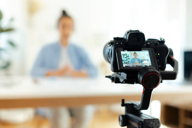 On a camera screen pretty indian or arabian brunette young woman, blogger, influencer sits at a desk, looking at camera, recording education tutorial vlog, video course for social media stock photo