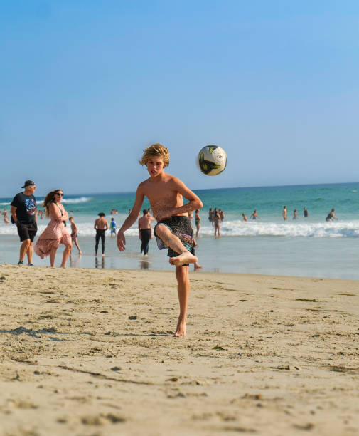 A young boy juggles a soccer ball effortlessly on the sandy shores of Monarch Beach at Laguna Niguel, n Orange County. The sun shines brightly overhead, illuminating his playful spirit as he showcases his skills against the backdrop of gentle waves and a clear blue sky.