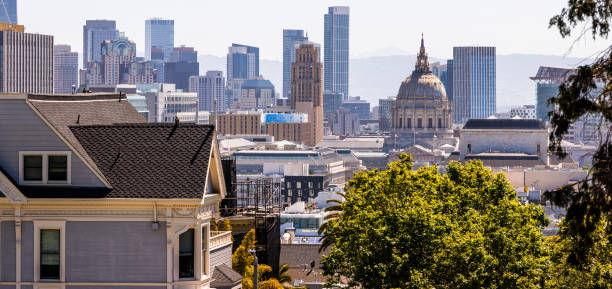 San Francisco's downtown skyline in morning light of California. City Hall dome stands out among modern skyscrapers. Traditional Victorian houses in the foreground contrast with the urban backdrop. Lush green trees frame the scene, showcasing the unique blend of historic charm and contemporary urban development defining San Francisco.