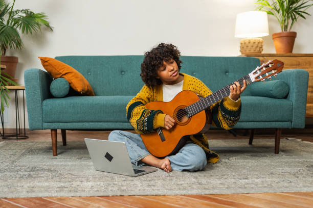 istock African American woman learning guitar watching tutorial on laptop at home. Black girl playing guitar singing song learning online music lessons, Artistic woman playing acoustic guitar learning chords 2176748616