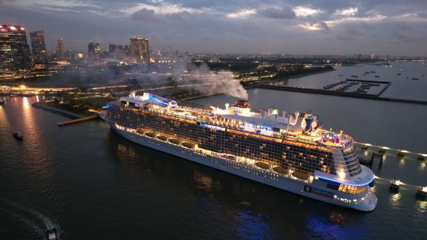 Staten Island, NY, USA - August 3, 2025: Symphony of the Seas cruise liner navigates the Narrows under the Verrazzano Bridge. The ship is adorned with multiple decks, numerous yellow lifeboats, and has several levels of balconies. The bridge features tall vertical towers and a series of suspension cables, creating an elegant arc over the water.