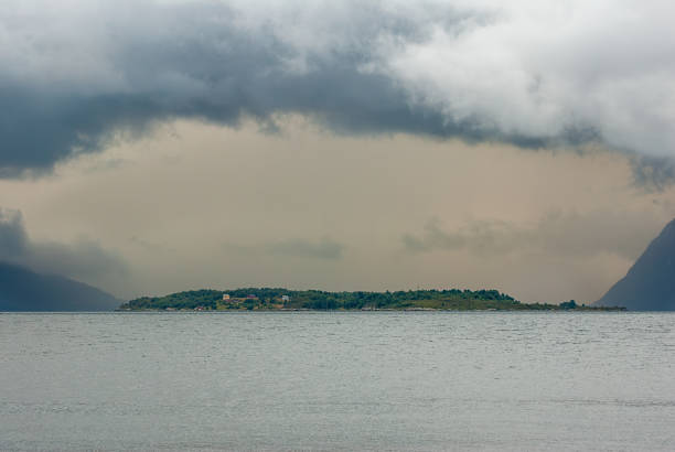 Amidst swirling clouds, an island reveals its lush greenery, quietly reflecting under the soft twilight on a fjord.