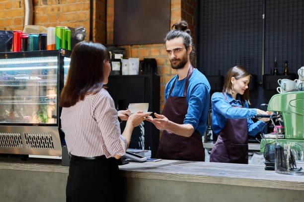 istock Guy working in cafe with cup of coffee at bar counter serving female client 2176871246