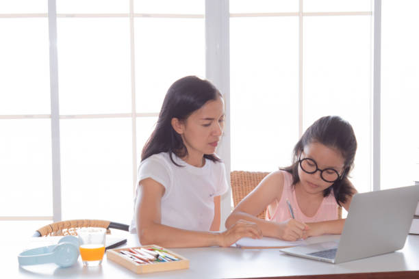 istock Asian single mother sitting with daughter, helping with homework while using laptop, emphasizing parent-child learning and guidance. Nanny helps primary school children with homework during holidays. 2176872345