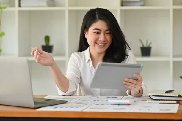 istock Portrait with Young Asian businesswoman using laptop and tablet while analyzing business graphs, diagrams and figures on financial document. 2176898771