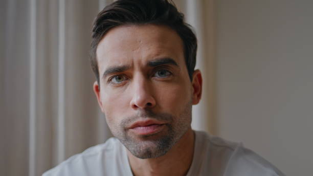 Closeup serious unshaven guy looking camera sitting apartment room alone. Portrait on pensive dark-haired man staring lens with tensed gaze. Thoughtful young male model posing at home interior alone.