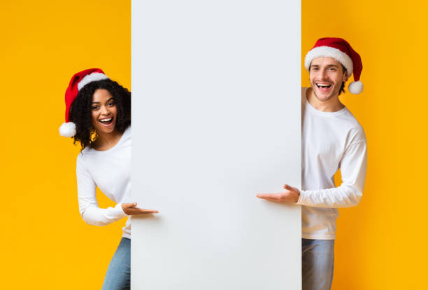 istock Cheerful Interracial Couple In Santa Hats Indicating At White Advertisement Board 2176906485
