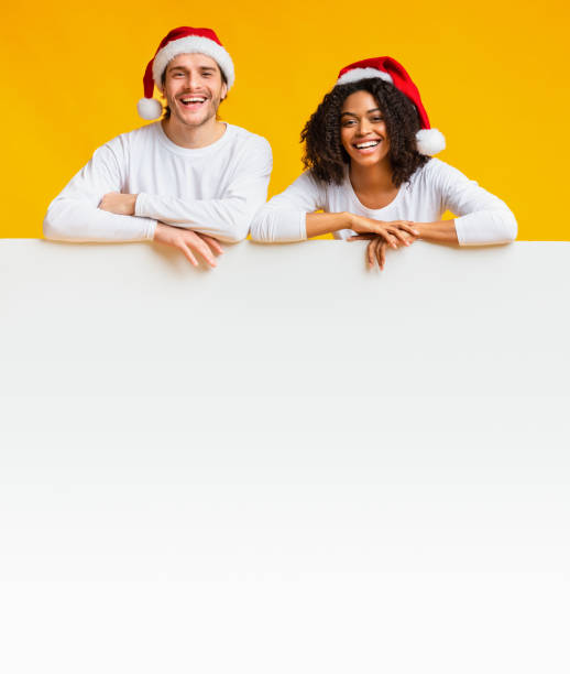 istock Interracial Couple In Santa Hats Leaning On White Blank Advertisement Board. 2176906500