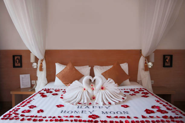 Shot of two folded swan towel decoration with a happy honeymoon text writing on top of a bed, in honeymoon room at villa in bali. The bed is covered by red rose petals. concept : hotel room decoration for honeymoon event.