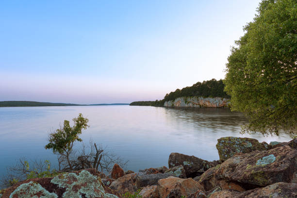 A serene dusk scene at Atoka Oklahoma Park, showcasing the calm lake waters reflecting the soft hues of the sky. The rocky shoreline and dense tree line create a peaceful and scenic environment perfect for nature lovers.