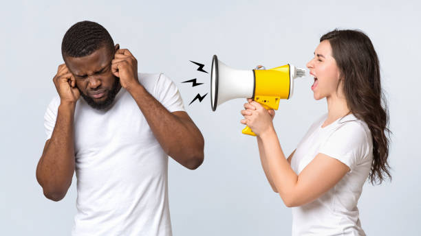 Angry woman shouting at her boyfriend with loudspeaker over light background Angry woman shouting at her afro boyfriend with loudspeaker, having nervous breakdown or pms, standing together over light background, panorama man closing ears photos stock pictures, royalty-free photos & images
