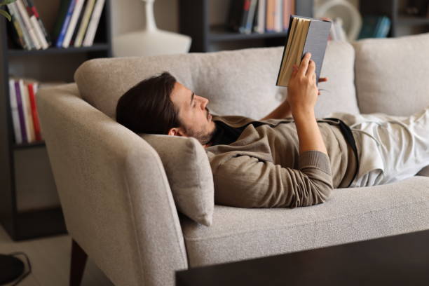 istock Young bearded handsome man reading book on couch at home 2176974819