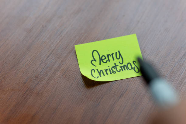 A person is writing on a sticky note on a table wood background. the person is writing Merry Christmas