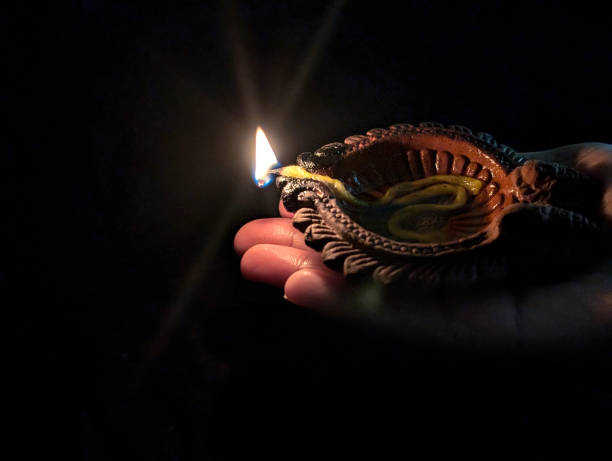 Lighted Shining illuminated terracotta clay crafted shubh Diwali Diya lamp with peaceful serene flame over dark black night horizontal backgrounds in side view of one hand for festive celebrations greeting card, banner and poster with copy space. Apt for backdrops, Greeting cards templates, posters, gift wrapping paper related to festivals Durga Pooja, Ganesh Chaturthi, Navratri and Indian prayers rituals.