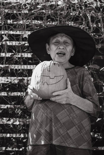 A kind Japanese elderly female happy celebrating Halloween the western way on her own, by wearing an improvised witch costume, carving and holding a Jack-O-Lantern bright orange Japanese pumpin.
