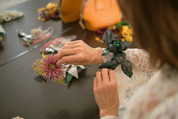 Hands delicately handle various floral accessories on a table, with an orange handbag in the background. The photo captures the creative and detailed process of choosing the right accessories.