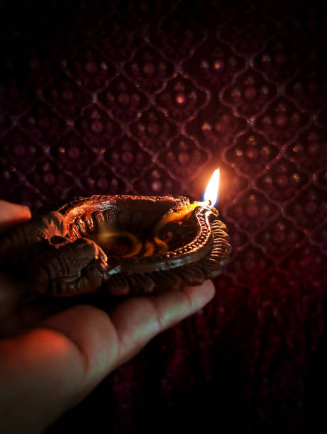 istock Offering Prayers with one lighted Shining illuminated terracotta clay shubh Deepawali, Diwali Diya lamp with peaceful serene flame over dark black night vertical backgrounds carried by or placed in one hand against ethnic motif backdrop 2177060915