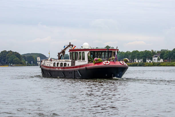 Red ship decorated with bouquets of flowers sailing on river Meuse, hills and leafy trees in background, landscape outside city of Maastricht in South Limburg, Netherlands