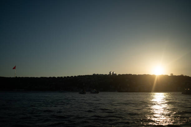 A breathtaking view of the Istanbul Bosphorus at sunset, featuring the iconic Turkish flag waving proudly. The golden light reflects on the water, highlighting the city's historic skyline and creating a serene, picturesque scene.