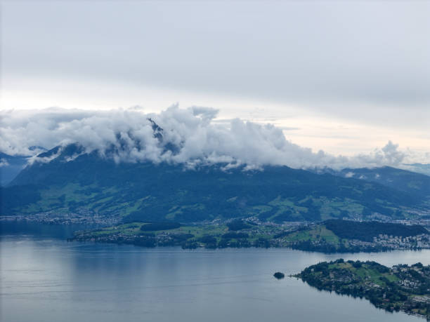 Scenic aerial view of a mountain partially covered by low clouds, with a tranquil lake and small villages nestled along the shoreline, creating a peaceful natural landscape.