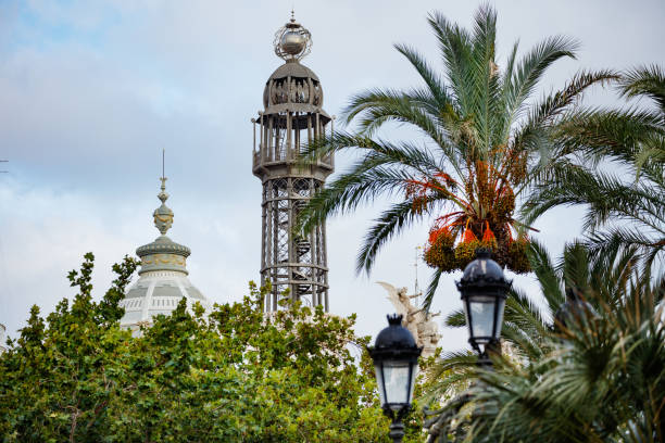 The steel Barcelona Port Cable Car Tower rises above palm trees and buildings under a clear blue sky.