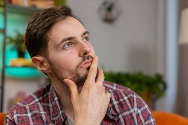 Thoughtful Caucasian man young guy sitting at home looks pensive thinks over life concerns or unrequited love, suffers from unfair situation. Problem, break up, depressed feeling bad annoyed, burnout